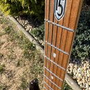 Detail hmatníku elektrické kytary s dřevěnou povrchovou vrstvou a kovovými pražci. Na hmatníku je umístěn dekorativní štít s číslem 5. Na dřevě jsou…