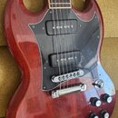 Elektrická kytara s červeným lakovaným povrchem s viditnou strukturou dřeva a černým pickguardem. Nástroj má dva humbuckery, stříbrný most a stop-bar…