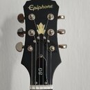 Fotografie zachycuje hlavu černé elektrické kytary. V horní části je bílý nápis Epiphone a uprostřed se nachází světle žlutý ornament ve tvaru koruny…