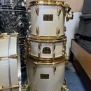 Tři bílé bubny s pozlaceným hardwarem naskládané na sebe. Na každém z nich je viditelný štítek s nápisem Pearl Master Custom. Povrch nástrojů a kovov…