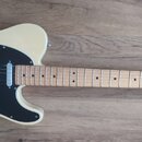 Elektrická kytara s krémovým korpusem, černým pickguardem a světle hnědým hmatníkem. Na hlavě krku je nápis Fender TELECASTER. Nástroj vykazuje čistý…