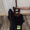 Elektrická kytara s tmavě hnědým sunburst povrchem, bílým pickguardem a hnedým hmatníkem. Na hlavě je viditelné logo Fender. Kytara je umístěna nad o…