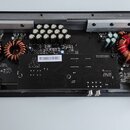 Fotografie znázorňuje vnitřek elektronického zařízení, pravděpodobně zesilovače, v černém plastovém šasi. Na černé desce plošných spojů je viditelné…