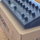 Černý syntezátor s nápisem ARGON8M položený na hnědé kartonové krabici, na které je vidět text ARGON8M a část loga MOOG ELECTRONICS. Přístroj má mnoh…