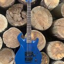 Modrá elektrická kytara s tmavým hmatníkem, umístěná na hromadě dřevěných polen. Na hlavě nástroje je svisle napsané logo "Ibanez". Kytara má dva hum…