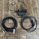 Na dřevěném podkladu jsou položeny tři předměty: černý síťový adaptér s přívodním kabelem, černý USB kabel a černý audio kabel. Audio kabel má na kon…