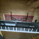 Černý MIDI kontrolér s klaviaturou, umístěný na černém X-stojanu. Na horním panelu je bílý nápis novation, sada padů a řada posuvníků. Povrch nástroj…