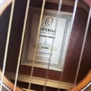Detail vnitřku akustické kytary s viditelným štítkem. Na štítku je logo a nápisy ORTEGA GUITARS • EST 1994, THERMO SERIES a v samostatném políčku ozn…