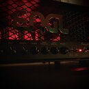 Kytarový zesilovač s černým povrchem a kovovou mřížkou, za kterou jsou vidět červeně osvětlené elektrony. Na mřížce je umístěno výrazné stříbrné logo…