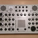 Bílý syntetizátor v dřevěném rámu s mnoha ovládacími knoflíky, přepínači a zapojovacími konektory. Na horní části je umístěn analogový VU metr s nápi…