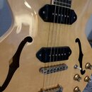 Detailní pohled na tělo polodupaté elektrické kytary v přirozené dřevěné barvě s lakovaným povrchem. Nástroj má dva černá humbuckery, chromovaný koby…