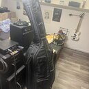Černý polstvany obal na hudební nástroj s nápisem G.Berton. Obal má několik zipových kapes a je opřen o černý kombozesilovač. Povrch materiálu vykazu…