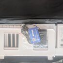Bílá elektronická klávesa v černé přepravní tašce. Na pravé straně je ovládací panel s digitálním displejem a nápisem DOHNER. Na nástroji leží napáje…