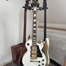 Na fotografii jsou tři elektrické kytary v držáku. V popředí je bílá kytara typu Les Paul s černým pickguardem a dvěma humbuckery, na hlavě má nápis…