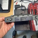 Ruka drží černý, obdélníkový elektronický přístroj s několika konektory a ovratkem. Na boku zařízení je viditelné logo ve tvaru stylizovaného písmene…