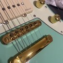Detail elektrické kytary v tyrkysové barvě s bílým pickguardem. Na snímači je viditelný nápis Roswell a kytara má zlaté hardware, včetně mostu a knob…