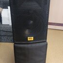 Černý reproduktor s kovovou mřížkou, na které je umístěn žlutý štítek s nápisem SPL Acoustics. Reproduktor stojí na černém polstrovaném podstavci, ve…