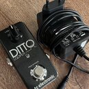 Černý kytarový pedál s nápisy DITTO LOOPER, TRUE BYPASS a tc electronic. Vedle pedálu je černý síťový adaptér s kabelem a evropskou vidlicí, na které…