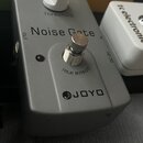 Šedý efekt pedál s nápisem Noise Gate a logem JOYO. Na horní části je bílý oválný knoflík s popiskem Threshold. Uspřádání ovládacích prvků doplňuje s…