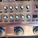 Kovový akustický preamp s nápisem Acoustic Preamp AD-10 a logem BOSS. Zařízení obsahuje řadu ovládacích potenciometrů, přepínačů a LED indikátorů roz…