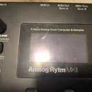 Detailní pohled na ovládací panel černého elektronického přístroje. Na zařízení je displej, nad ním nápis „8 Voice Analog Drum Computer & Sampler“ a…
