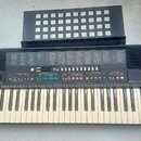 Černý elektronický klávesový nástroj s bílými a černými klávesami. Na levé straně je bílé logo YAMAHA a na pravé straně nápis PSR-215. Nad klávesami…