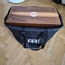 Hnědý dřevěný cajón s logem MEINL umístěným uprostřed horní plochy. Nástroj je vložen do černé látkové přepravní tašky, která má na přední straně bíl…