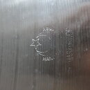 Detail kovového povrchu s broušenou strukturou. Je na něm vyryto logo ve tvaru půlměsíce s hvězdou a nápisy THEY a HAND. Vedle loga je svisle vyryto…