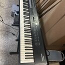 Černý digitální klavír na černém stojanu. Na pravé straně korpusu je bílý nápis „Roland RD-2000“. Nástroj vykazuje běžné známky používání a je umístě…