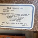 Fotografie detailně zachycuje informační štítek na povrchu z dřevotřisky. Na štítku je černý text: LESLIE TREMOLO UNIT, PART NO. 79019, EMI NO., MANU…