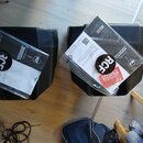 Dvě černé reproduktorové bedny s plastovým povrchem. Na každé z nich leží v plastovém obalu záruční list s nápisy „2 YEARS WARRANTY CERTIFICATE“ a lo…