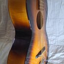 Akustická kytara s tělem v barvě sunburst (přechod od žluté přes oranžovou k hnědé). Nástroj má tmavý hmatník a černý kobylník. Na viditelné části ná…