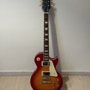 Elektrická kytara s trupem v červeně-žlutém sunburst provedení a bílým pickguardem. Na hlavě hmatníku je bílý nápis Epiphone a pod ním kurzivou Les P…