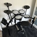 Elektronická bicí souprava s černým rámem a černými činely. Na bubnech je vidět logo Roland, na sedátku nápis Pearl. Souprava obsahuje pět padů, tři…