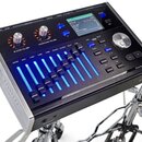 Černý elektronický bicí modul s kovovým bočním rámem. Na předním panelu jsou dva otočné ovladače s nápisy MASTER VOLUME a HEADPHONE VOLUME, řada modr…
