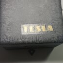 Černý hranatý box s povrchem z texturovaného materiálu připomínajícího kůži. Uprostřed je umístěn zlatý nápis TESLA. Povrch vykazuje mírné opotřebení…