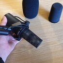 Černý dynamický mikrofon s mřížkou, na kterém je bílý nápis SHURE SM7B. Mikrofon je upevněn v černém držáku a drží ho ruka. Vedle mikrofonu leží dvě…