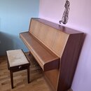 Dřevěný pianino v hnědé barvě s doprovodnou stolečkou s béžovým čalouněním. Nástroj je umístěn v rohu místnosti u růžové a modré stěny. Na povrchu ná…