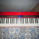 Červený digitální klavír s bílými a černými klávesami. Na levé straně ovládacího panelu je logo Roland a na pravé straně nápis GO:KEYS. Uprostřed se…