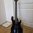 Černá elektrická kytara s dvojsekmem, umístěná na černém stojanu. Na hlavě nástroje je bílý nápis CORT. Kytara má dva humbuckery a dva ovládací knofl…