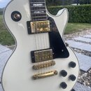 Bílá elektrická kytara s černým pickguardem a zlatými komponenty. Na těle jsou dva humbuckery, most a strunník v zlatém provedení a čtyři černé oválk…