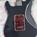 Zadní strana elektrické kytary v tmavě šedé barvě s lesklým povrchem. Na spojení krku a těla je kovovádeska s nápisem Fender STRATOCASTER. V dolní čá…
