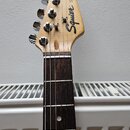 Fotografie zachycuje hmatník a hlavu elektrické kytary z přírodního dřeva s tmavým hmatníkem. Na hlavě je černé logo Squier a nápis STRAT®. Povrch ná…