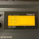 Detailní pohled na ovládací panel šedého elektronického modulu značky Roland. Na displeji je zobrazen text 01 001 Piano 1, LEVEL 100 a PAN L46. Pod d…