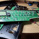Fotografie zachycuje rozebraný elektronický hudební přístroj na stole. V centru je zelená základní deska s LCD displejem, několika potenciometry a za…