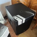 Černý subwoofer v hranatém korpusu s texturovaným povrchem. Přední strana je kryta černou kovovou mřížkou, v jejím spodním okraji je umístěn malý neč…