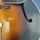 Detailní pohled na horní desku akustické kytary s f-otvorem. Nástroj má tmavě hnědý přechodový lak (sunburst) a je vybaven plektrem. Povrch vykazuje…