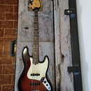 Elektrická basová kytara v tvrdém kufru s šedým polstrováním. Nástroj má korpus s tmavě hnědým sunburst povrchem, bílý pickguard a hmatník z tmavého…