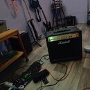 Na fotografii je kytarový kombozesilovač značky Marshall v černé barvě se zlatým ovládacím panelem. V pozadí jsou vidět tři elektrické kytary, stojan…