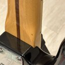 Detail krku a spoje s tělem elektrické kytary. Krk je vyroben ze světlého dřeva s tmavým středovým pruhem, tělo má černý lesklý lak. Na kovém krytu s…
